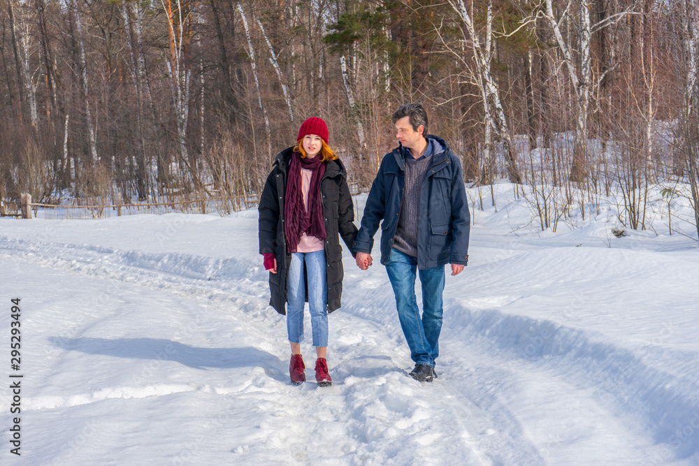 Handsome man and attractive young woman walking along snowy country road in sunny day. Beautiful look, male and female fashion, winter outfit. Winter holidays, weekend at countryside concept