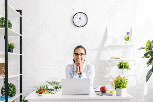cheerful nutritionist in white coat near vegetables and laptop photo