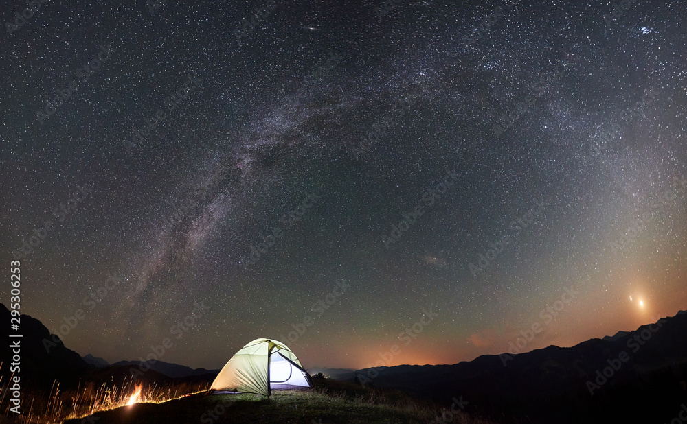 Panoramic view of tourist camping at night in the mountains. Illuminated tent and campfire under beautiful night sky full of stars and Milky way