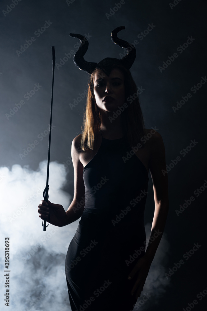 Foto de silhouette of young woman with horns holding flogging whip on black  with smoke do Stock