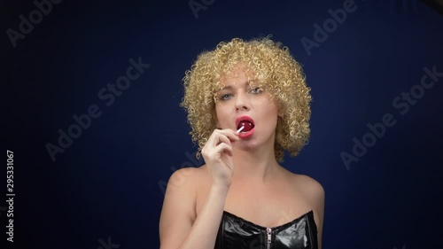 sweet Woman with short curly blond hair in a black leather corset licks a red shiny lollipop. dark background photo