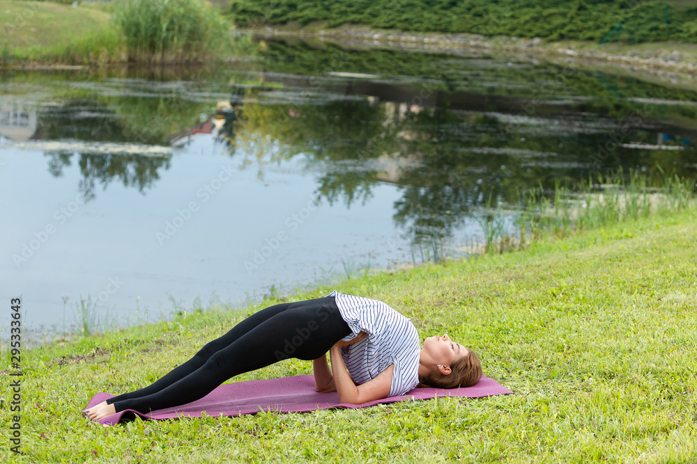 Fototapeta premium Young beautiful woman doing yoga exercise in green park near the pond. Exercices for improve the flexibility. Wellbeing and wellness. Healthy lifestyle and fitness concept.