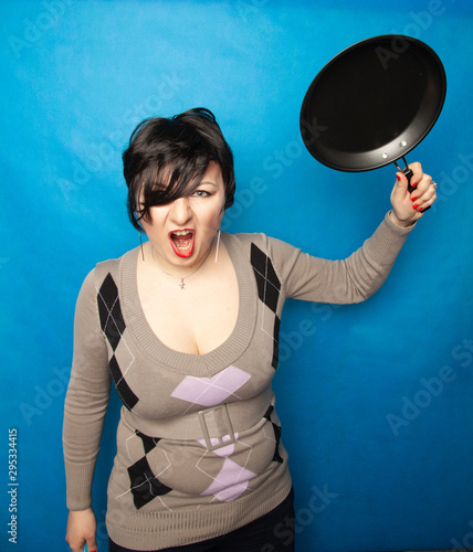 Woman beating with a frying pan on bluw studio background alone photo