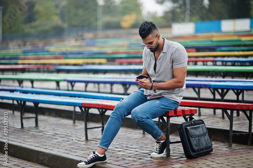 Fashionable tall arab beard man wear on shirt, jeans and sunglasses sitting on colored row of benches.