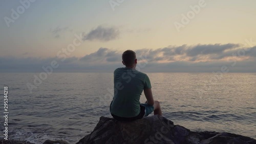 A man sits at sunset on a large stone and relaxes. Rear view silhouette of a man at sunset.