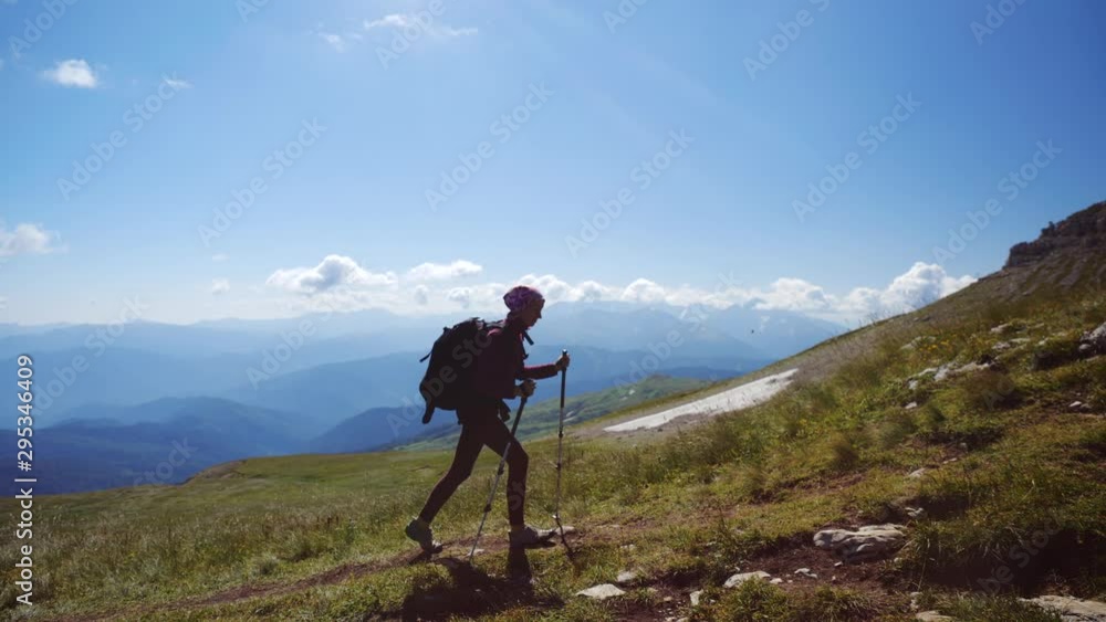 Young woman with backpack and trekking poles walks uphill towards the summit against blue sky. Slow motion. Lady is hiking in beautiful Caucasian mountains. Young woman with backpack and trekking poles walks uphill towards the summit against blue sky. Slow motion. Lady is hiking in beautiful Caucasian mountains.