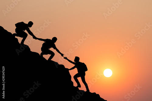 Silhouette of people helping each other hike up a mountain at sunset background. Teamwork, success and goal concept.