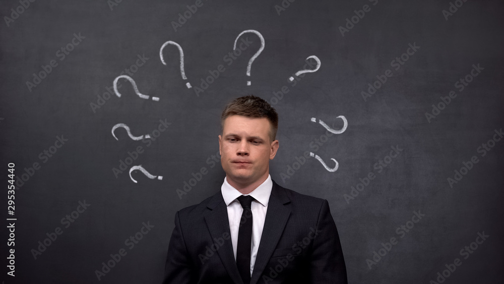 Doubtful businessman lacking ideas, question marks painted on blackboard, answer Doubtful businessman lacking ideas, question marks painted on blackboard, answer