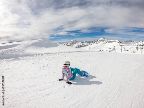 A snowboarder sitting on the slope in Mölltaler Gletscher, Austria. Perfectly groomed slopes. High mountains surrounding the girl wearing colourful snowboard outfit. Girl wears helm for the protection