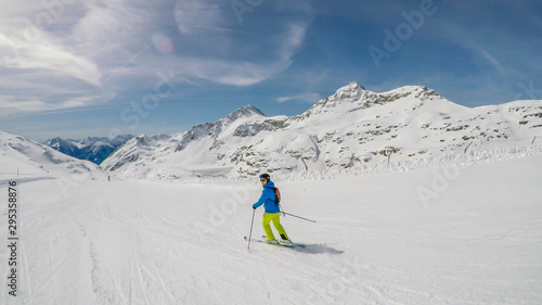 A skier going down the slope in Mölltaler Gletscher, Austria. Perfectly groomed slopes. High mountains surrounding the man wearing yellow trousers and blue jacket. Man wears helm for the protection.