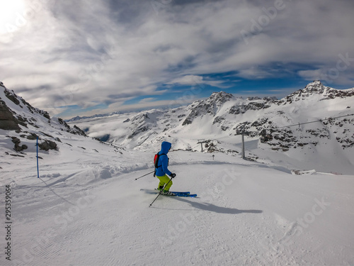 A skier going down the slope in Mölltaler Gletscher, Austria. Perfectly groomed slopes. High mountains surrounding the man wearing yellow trousers and blue jacket. Man wears helm for the protection.