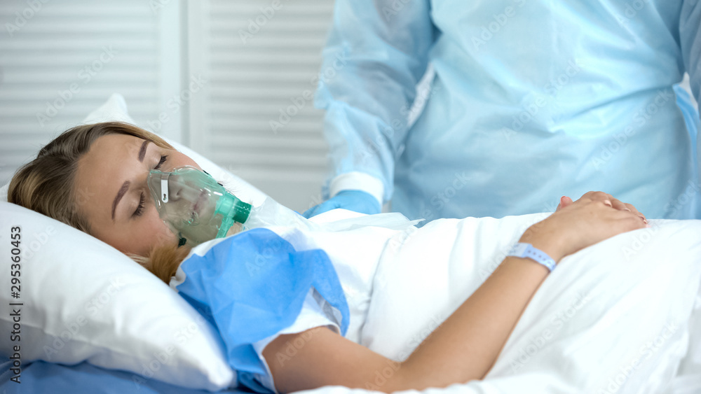Fototapeta premium Female patient in oxygen mask sleeping, nurse standing by, surgery preparation