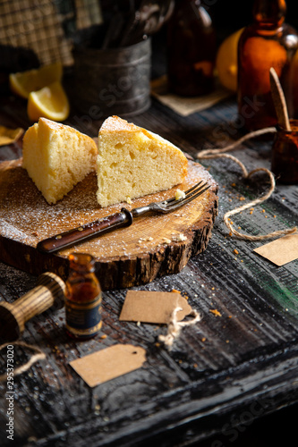 homemade two tasty slices of baked lemon biscuit cake with powdered sugar on top stands on wooden board on rustic table with lemons, old bottles opposite concrete wall, selective focus