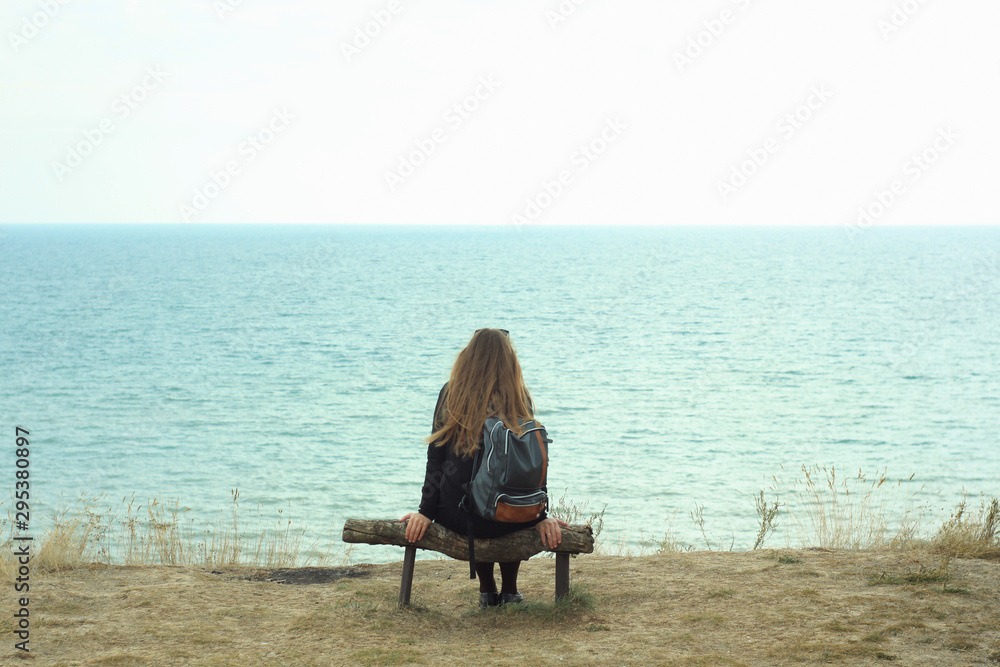 A young woman travels alone. Solo travelling. A tourist sits on a wooden bench on a cliff above the sea coast and looks at the horizon. Loneliness..