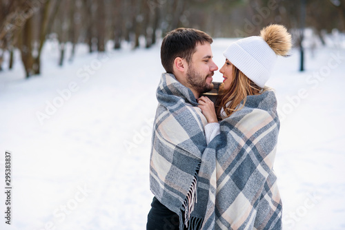Beautiful couple hugging each other and covered with checkered plaid while walking in snowy winter park. © gorynvd