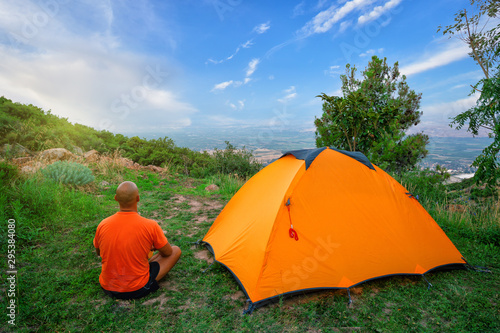 Man in an orange T-shirt sits near tent on hill