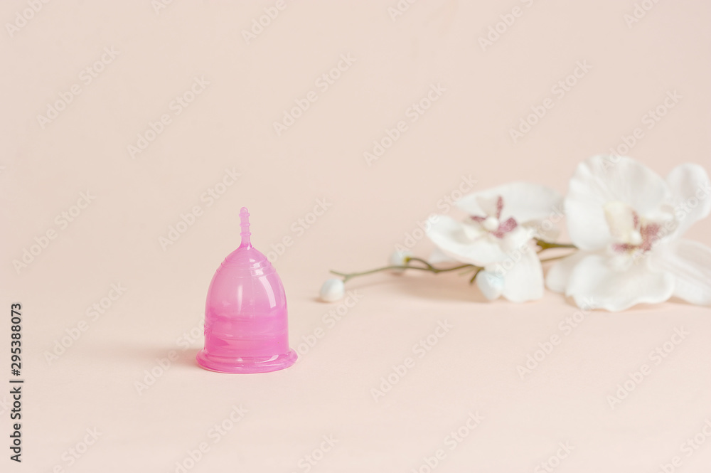 Pink menstrual cup in the shape of a lily on a light background. Nearby is an orchid flower. The concept of cleanliness, comfort and freshness. Free space for text. Pink menstrual cup in the shape of a lily on a light background. Nearby is an orchid flower. The concept of cleanliness, comfort and freshness. Free space for text.