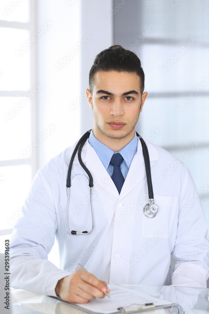 Arab doctor man using laptop computer while filling up medication history records form at the glass desk in medical office or clinic. Medicine and healthcare concept Arab doctor man using laptop computer while filling up medication history records form at the glass desk in medical office or clinic. Medicine and healthcare concept