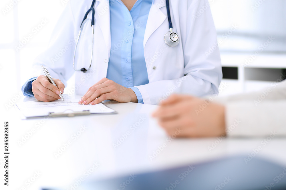 Woman doctor and patient sitting and talking at medical examination at hospital office, close-up. Physician filling up medication history records. Medicine and healthcare concept Woman doctor and patient sitting and talking at medical examination at hospital office, close-up. Physician filling up medication history records. Medicine and healthcare concept