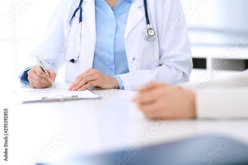 Woman doctor and patient sitting and talking at medical examination at hospital office, close-up. Physician filling up medication history records. Medicine and healthcare concept