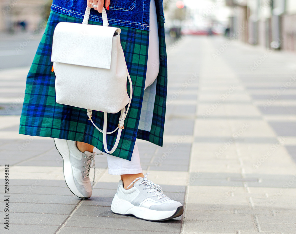 Fashionable bag close-up in female hands.Girl walks in the city outdoors. Stylish modern and feminine image, style. A woman in a raincoat, coat and with a white backpack. girl in sneakers and jeans. - obrazy, fototapety, plakaty 