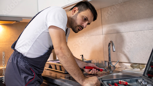 Plumber man fixing kitchen sink. Handsome plumber at the kitchen. Reparation. Handsome plumber repairing sink pipes in kitchen. serious male plumber in working overall fixing sink in kitchen