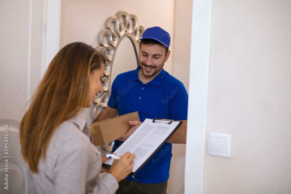 Woman Signing For Package From Courier At Home. Smiling young delivery man holding a cardboard box while beautiful young woman putting signature in clipboard Woman Signing For Package From Courier At Home. Smiling young delivery man holding a cardboard box while beautiful young woman putting signature in clipboard