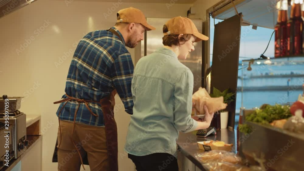 Street Food Joint Female Employee Hands Out Beef Burgers to Happy Hipster Customers. Male Employee is Preparing Food. Commercial Kiosk is Selling Street Food in a Modern Place Near the Sea. Street Food Joint Female Employee Hands Out Beef Burgers to Happy Hipster Customers. Male Employee is Preparing Food. Commercial Kiosk is Selling Street Food in a Modern Place Near the Sea.