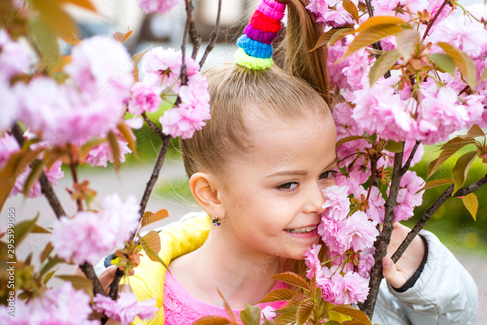 Fototapeta premium Smiling blonde girl standing in a blooming garden. Blooming cherry. Portrait of beautiful little girl. Close up of little model face. Sakura blooming, spring time.
