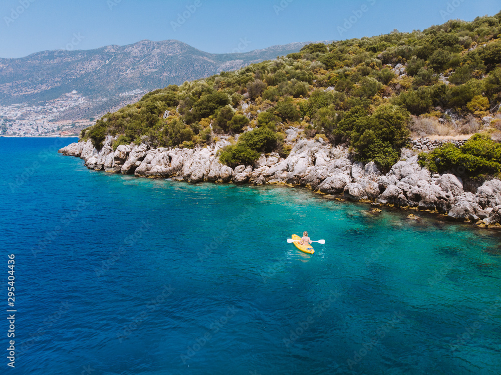 Fototapeta premium A lone girl paddling a yellow kayak in remote mediterranean waters, exploring the Turkish coastline. Beautiful blue water and coastline with amazing visibility. Shot aerially with a drone.