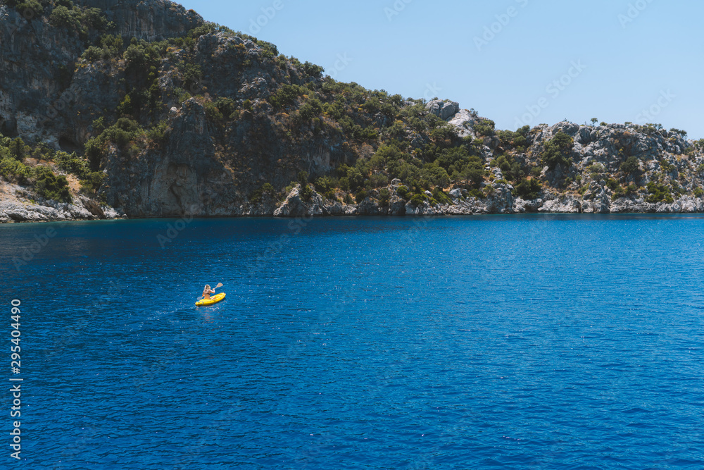 A lone girl paddling a yellow kayak in remote mediterranean waters, exploring the Turkish coastline. Beautiful blue water and coastline with amazing visibility. Shot aerially with a drone.