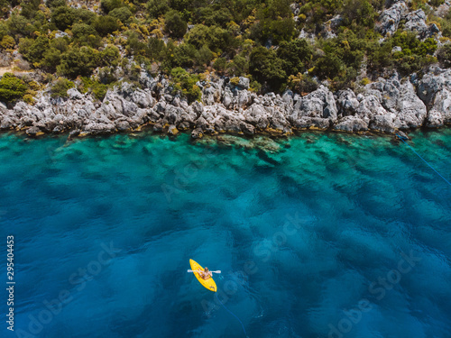 A lone girl paddling a yellow kayak in remote mediterranean waters, exploring the Turkish coastline. Beautiful blue water and coastline with amazing visibility. Shot aerially with a drone.