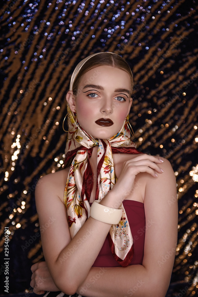 Fashion girl in a textured satin headscarf on head on a gold glitter blurred background. Professional evening make-up, brown lipstick, eyelid art, French manicure. Gold round earrings in the ears.