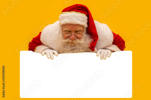 Happy Santa Claus looking out from behind the blank sign isolated on yellow background with copy space © Brastock Images