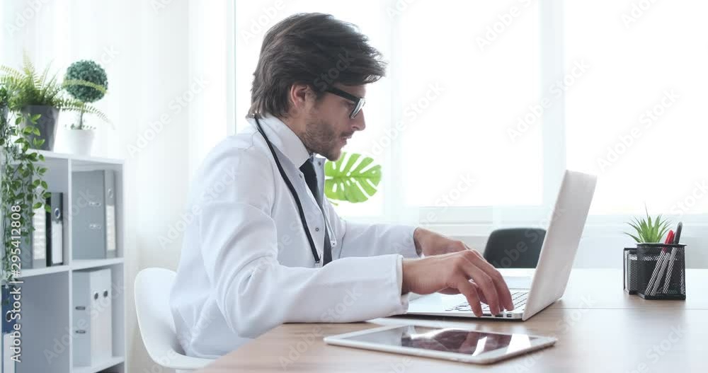 Doctor writing notes while examining xray report and working on laptop at clinic Doctor writing notes while examining xray report and working on laptop at clinic