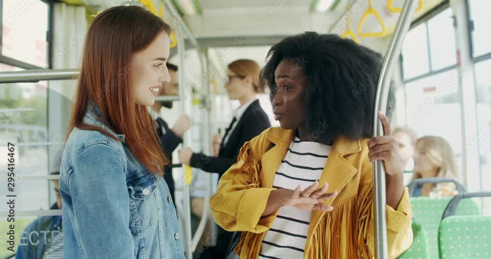 Two mixed-races young beautiful and joyful friendly girls standing in the tram and talking while going somewhere. Two mixed-races young beautiful and joyful friendly girls standing in the tram and talking while going somewhere.