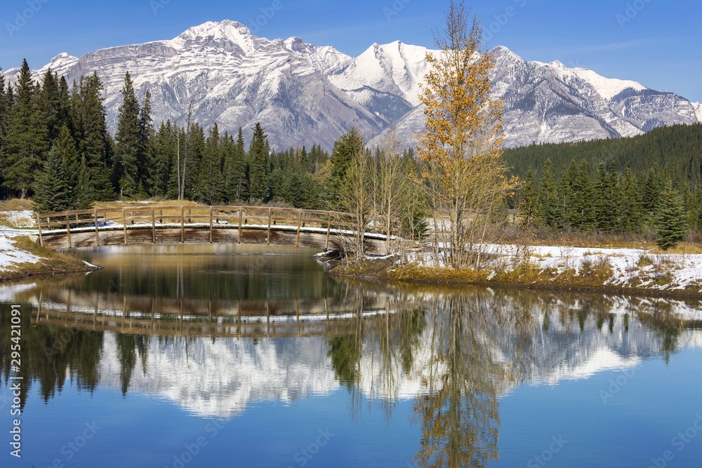 Pedestrian Hiking Footbridge Reflected in Calm Blue Water of Cascade Pond or Lake with Distant Snowcapped Mountain Peaks in Background. Banff National Park, Alberta Canada