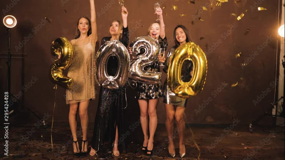 Beautiful Women Celebrating New Year Eve Party.Happy Gorgeous Girls In Stylish Dresses Holding Gold,Silver 2020 Balloons,Having Fun At Eve Party.They drinking champagne flutes under flying confetti