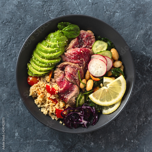 Healthy food buddha bowl with beef steak, beans, couscous, avocado and vegetables on dark background with copy space photo