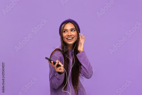 Pretty african-american girl in a hat and sweater listening to music on a mobile phone with wired headphones and dancing isolated over purple background. Enjoying life.
