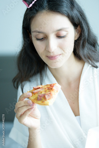 A beautiful woman wearing a towel and a white bathrobe has to eat a pizza with happy and relaxing on the bed at a condominium in the morning.