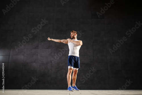 Full length of young muscular caucasian man in shorts and t-shirt doing warm up exercises for arms. In background is gray wall.