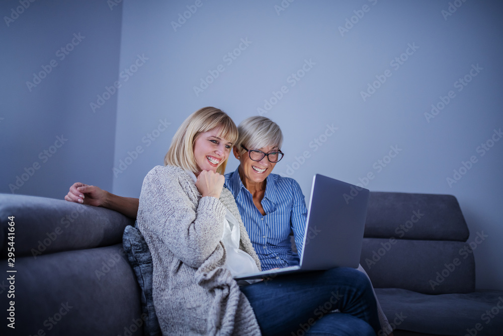 Smiling pregnant blonde woman sitting on sofa in living room and holding laptop in lap. Next to her is sitting her mother. Both are looking at laptop and smiling. Smiling pregnant blonde woman sitting on sofa in living room and holding laptop in lap. Next to her is sitting her mother. Both are looking at laptop and smiling.