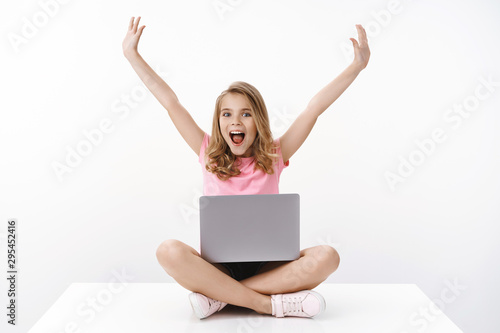 Happy cheerful cute blond girl child having fun spend summer holidays away from school, enjoy watching cartoons, sit crossed legs using laptop, excited studying at home with e-learning program © Cookie Studio