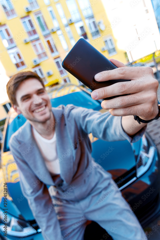 Closeup male hand is holding smartphone. Happy brunette man owner is making selfie with new car. Black automobile on parking on background. Smiling businessman in grey suit bought auto.