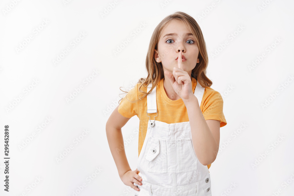 Keep quiet please. Determined obedient cute blond girl child showing hush sign, shushing asking silence, hold finger on lips turn off loud music, sharing secret, stand white background Keep quiet please. Determined obedient cute blond girl child showing hush sign, shushing asking silence, hold finger on lips turn off loud music, sharing secret, stand white background