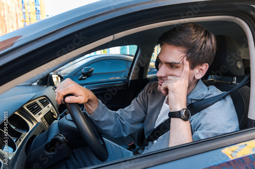 Annoyed tired young man is riding driving car. Businessman is late for meeting. Driver brunette in grey suit stuck in traffic jam. Stressful situations on roads and fast rhythm in modern city.