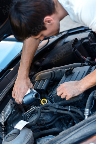 Young brunette man is changing oil in auto with open hood. Driver is repairing automobile on street road. Vehicle breakdown on way concept. Repairman mechanic is servicing car in workshop. © Monstar Studio