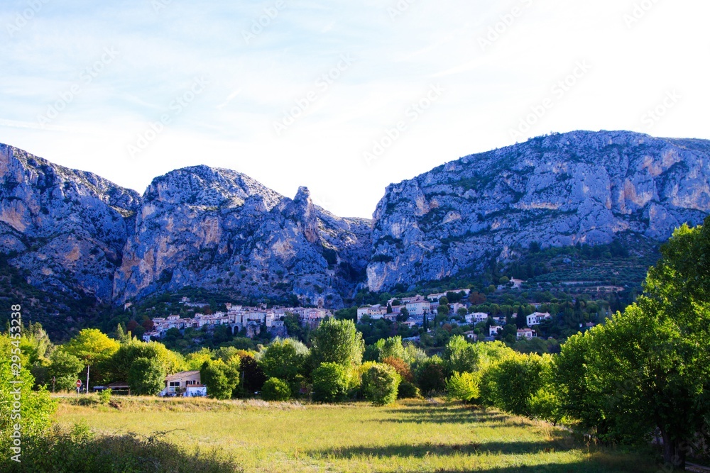 View over green valley on French village with giant mountain face background in early morning - Moustiers Sante Marie, Provence, France
