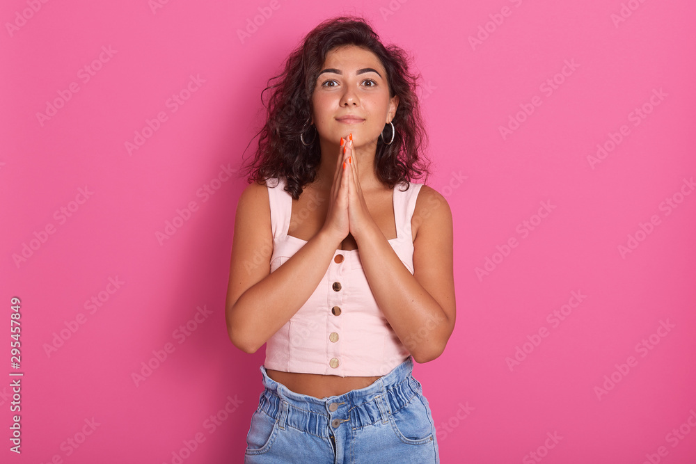 Close up portrait of pleasant looking glad Caucasian woman looking directly at camera, keeps hands together under chin, dreams about something, praying, wearing casual clothing, isolated on pink wall. Close up portrait of pleasant looking glad Caucasian woman looking directly at camera, keeps hands together under chin, dreams about something, praying, wearing casual clothing, isolated on pink wall.